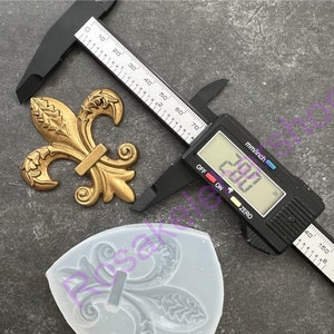 May include: A gold-colored metal fleur-de-lis design with a silicone mold of the same design. A digital caliper measures the metal design at 2.00 inches.