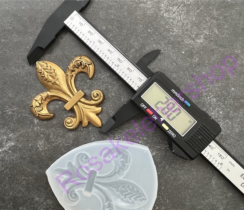 May include: A gold-colored metal fleur-de-lis design with a silicone mold of the same design. A digital caliper measures the metal design at 2.00 inches.