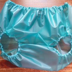 Puede incluir: Un pantalón de plástico turquesa con cintura y aberturas de piernas elásticas. El material brillante e impermeable está fruncido en la cintura y las aberturas de las piernas. Diseñado para la protección contra la incontinencia.