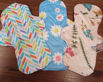 Puede incluir: Tres compresas menstruales de tela reutilizables con diferentes patrones. Una compresa tiene un fondo azul con margaritas blancas, otra tiene un fondo blanco con un patrón de chevron colorido y la tercera tiene un fondo rosa con hojas verdes y mariposas.