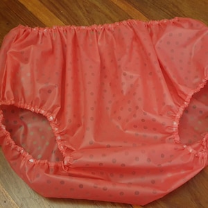 Puede incluir: Un pañal impermeable de color rosa con lunares blancos. El pañal tiene elástico alrededor de las piernas y la cintura.
