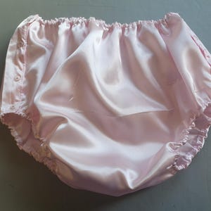 Puede incluir: Bragas de satén rosa claro con cintura y aberturas de las piernas elásticas. Las bragas tienen una cintura fruncida y se aseguran con botones en los laterales. La tela tiene una textura brillante y suave.