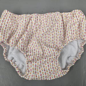 Puede incluir: Pañal de tela blanco con un estampado floral de pequeñas flores naranjas y rosas. El interior está forrado con un material blanco suave y absorbente. El pañal tiene elásticos alrededor de las piernas y la cintura.