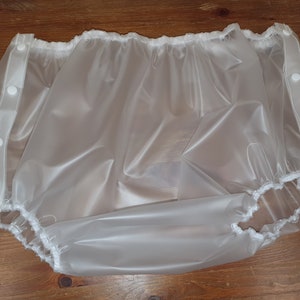 Puede incluir: Un pañal de plástico blanco con aberturas elásticas para las piernas y cierres a presión en los lados.