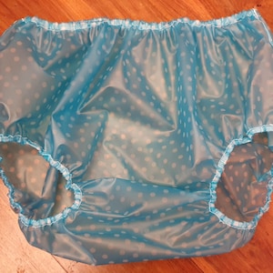 Puede incluir: Un pantalón de plástico azul claro con lunares blancos. El pantalón tiene cintura y aberturas de las piernas elásticas, diseñado para la protección contra la incontinencia. El material parece brillante e impermeable.