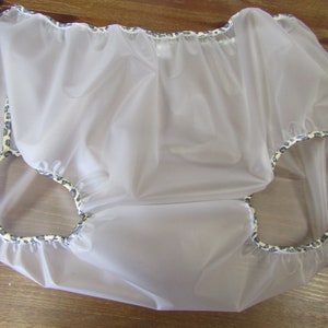 Puede incluir: Un pañal de plástico transparente blanco con un borde de estampado de leopardo. El pañal está hecho de un material suave y flexible y tiene una cintura elástica fruncida.