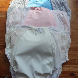 Puede incluir: Cuatro pares de bragas en blanco, rosa, azul y gris, cada uno con un panel frontal de satén y ribete de encaje. Pequeños lazos adornan la parte delantera de cada par. Las prendas están dispuestas sobre una superficie de madera.