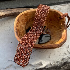 Peut inclure: Un bracelet en maille de chaîne en cuivre dans un bol en bois avec des pierres noires. Le bracelet est composé de petits anneaux interconnectés.
