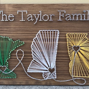 May include: String art of three states, Minnesota, Alabama, and Georgia, with a heart in each state. The states are connected by a string that forms a heart. The background is a brown wooden board. The text "The Taylor Family" is at the top of the board.