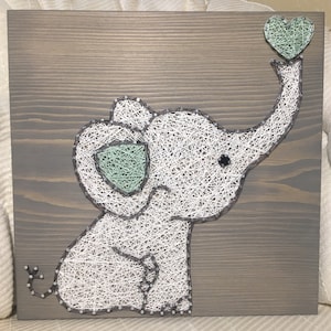 May include: A white string art elephant with a green heart in its trunk, on a brown wooden background.