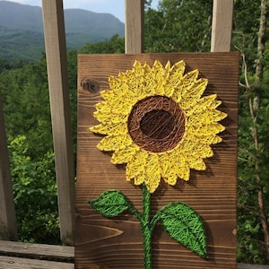 May include: A string art sunflower with a brown center and yellow petals on a wooden background. The sunflower has a green stem and two leaves.