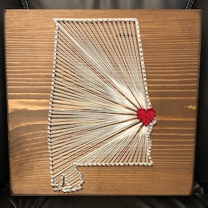 May include: String art of the state of Alabama with a red heart in the center. The art is on a wooden background.