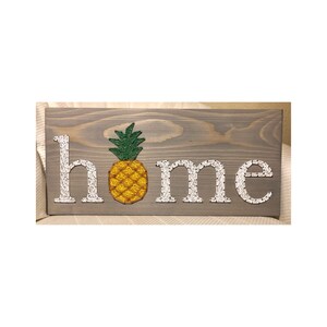 May include: A wooden sign with the word "home" spelled out in white string art. A pineapple is in the center of the sign, with green leaves and a yellow body.