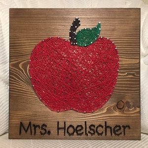 May include: A red apple string art piece on a wooden background with the text "Mrs. Hoelscher" below the apple.
