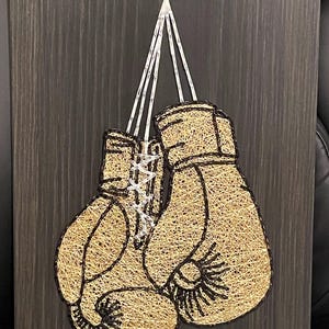 May include: A string art piece featuring two boxing gloves. The gloves are crafted with gold-colored string, with black detailing and white string laces. The artwork is mounted on a dark gray wooden board, creating a striking contrast.