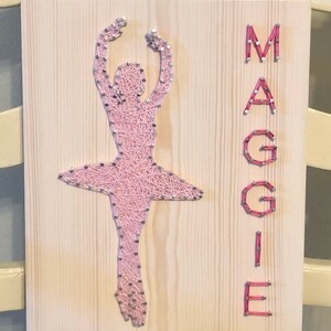 May include: A pink string art ballerina on a wooden background with the name "MAGGIE" spelled out in pink string art.
