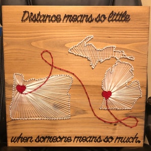May include: A wooden sign with the text "Distance means so little when someone means so much."  The sign features a string art design of the states of Michigan and Oregon connected by a red string with hearts at the center of each state.