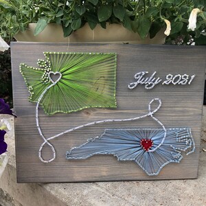 May include: String art of the states of Washington and North Carolina connected by a line with a heart in the middle. The states are outlined in green and blue thread on a wooden background. The text "July 2021" is written in white thread on the right side of the image.