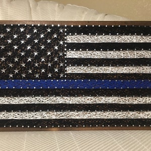 May include: A wooden framed string art piece depicting the American flag with a blue line running horizontally across the middle of the flag.