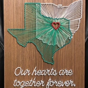 May include: A string art piece on a rectangular wooden board featuring the outlines of Texas and Ohio in green and white thread. A small red heart is positioned in the center. The text "Our hearts are together forever." is written in white string.