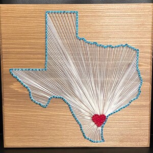 May include: A wooden wall art piece featuring a string art design of the state of Texas. The outline is in turquoise, with white string radiating from a red heart in the center. The wooden background has a natural wood grain.