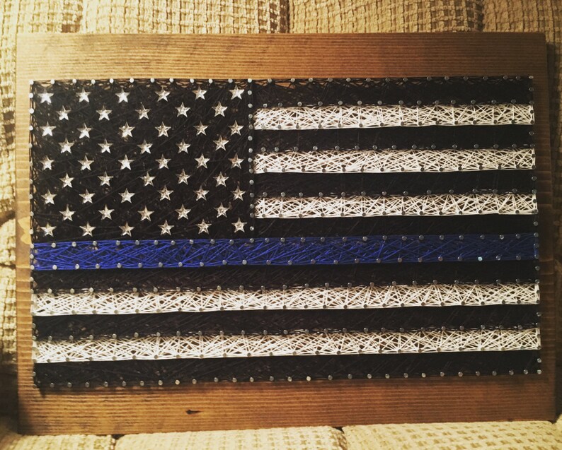 May include: A string art American flag with a blue line running horizontally across the middle of the flag. The flag is made of white and black string wrapped around nails hammered into a wooden board.