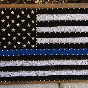 May include: A wooden framed string art piece depicting the American flag with a blue line, representing support for law enforcement.