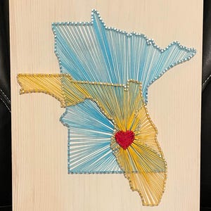 May include: A string art piece on a light wood background. The artwork depicts the states of Florida and Minnesota, with blue and yellow thread outlining the shapes. A red heart is centered in Florida.