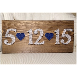 May include: A wooden sign with the date 5-12-15 created with white string wrapped around nails. Two blue hearts separate the numbers.