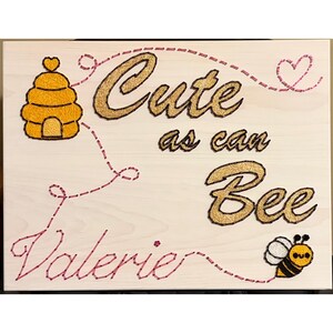 May include: Wooden sign with the text "Cute as can Bee Valerie" in gold glitter lettering and a cartoon bee and beehive. The sign has a light wood background with pink stitching accents.
