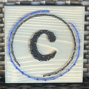 May include: A wooden square sign with a black string art design of the letter "C" surrounded by two concentric circles of blue and purple string with beads.