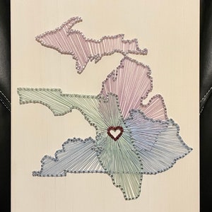 May include: A string art piece depicting the outline of the state of Michigan, with a heart in the center. The artwork uses various colors of thread, including purple, green, and blue, against a light-colored wooden background.