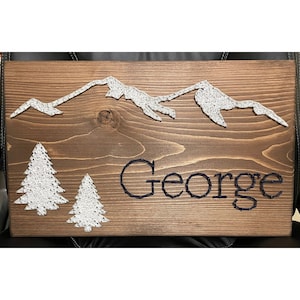 May include: A wooden sign with a mountain range and two pine trees made of white string art. The name "George" is spelled out in blue string art.