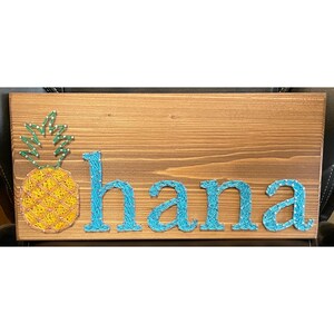 May include: A wooden sign with the word "ohana" spelled out in blue string art. A pineapple is depicted in the upper left corner of the sign.