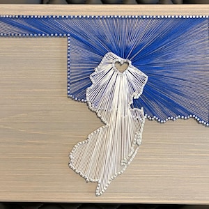May include: A string art piece on a light gray wooden board featuring the outline of Oklahoma in blue thread and the outline of New Jersey in white thread. The New Jersey section includes a small heart-shaped cutout near the top center.