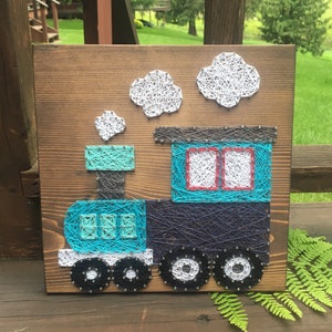 May include: A wooden sign with a blue and white string art train design. The train has a blue engine with a gray chimney and white smoke. The train has three wheels and a blue caboose with two windows.