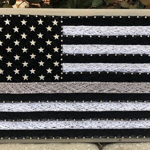 May include: A black and white string art American flag with a thin white stripe across the top, representing the thin blue line. The flag is framed in a light gray wood frame.