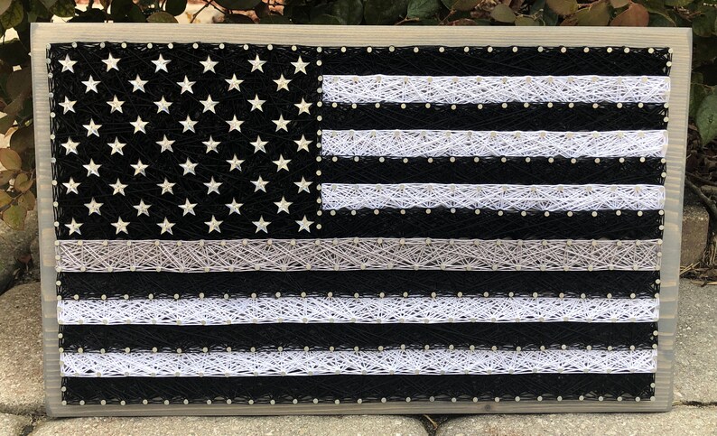 May include: A black and white string art American flag with a thin white stripe across the top, representing the thin blue line. The flag is framed in a light gray wood frame.