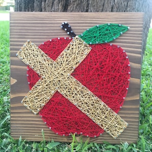 May include: A red apple with a gold X over it, made with string art on a wooden background.