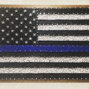 May include: A string art rendition of the American flag with a blue line running horizontally across the middle. The flag is made of white and black string wrapped around nails hammered into a wooden frame.