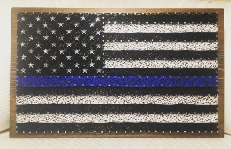 May include: A string art rendition of the American flag with a blue line running horizontally across the middle. The flag is made of white and black string wrapped around nails hammered into a wooden frame.