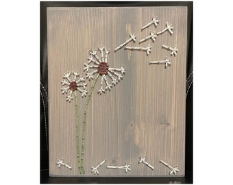 May include: A wooden wall art piece featuring two white dandelions with brown centers and green stems. The dandelions are surrounded by smaller white dandelions with brown centers, which are scattered around the piece.