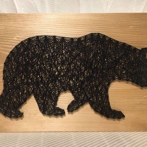 May include: Black bear silhouette created with string art on a wooden board. The bear is facing right and walking.