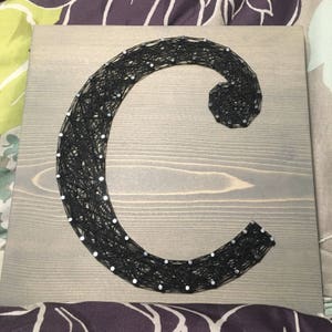 May include: A black string art piece in the shape of the letter C on a gray wooden background.