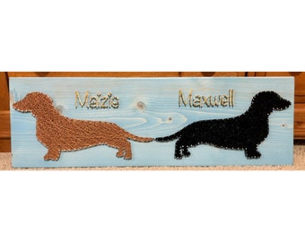 May include: A blue wooden sign with two dachshund silhouettes made of string art. The dachshund on the left is brown and says "Meizie" in gold lettering. The dachshund on the right is black and says "Maxwell" in gold lettering.