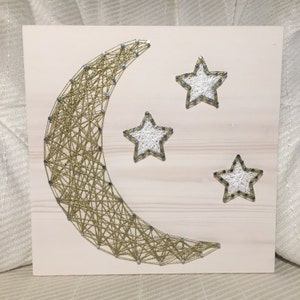 May include: A gold crescent moon and three white stars are created with string art on a light wood background.
