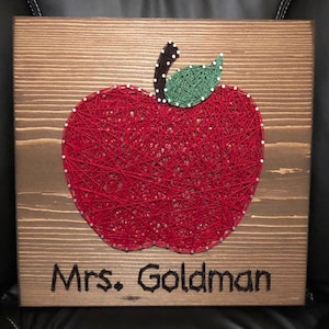 May include: A square wooden plaque featuring string art of a red apple with a green leaf and a brown stem, created with silver nails. Below the apple, the name Mrs. Goldman is spelled out in black string art, centered on the stained wood surface.