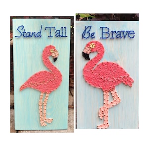 May include: Two wooden signs with string art of pink flamingos. The sign on the left says "Stand Tall" and the sign on the right says "Be Brave".