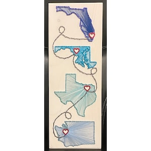 May include: A wooden string art piece featuring outlines of the states of Florida, Maryland, Texas, and Washington. Each state is crafted with blue string and connected by a gray string, with a red heart marking a location in each state.