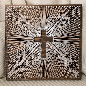 May include: A white string art cross on a brown wooden background. The cross is made of white string wrapped around nails hammered into the wood.